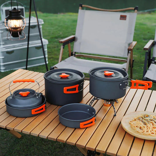 Camping cookware set with orange handles on outdoor table, chairs and lantern nearby