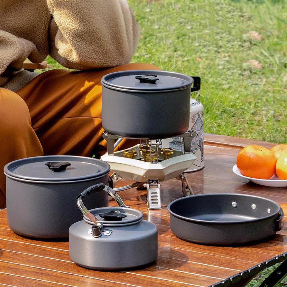 Camping cookware set with pots, pan, and kettle on wooden table outdoors, ideal for outdoor cooking